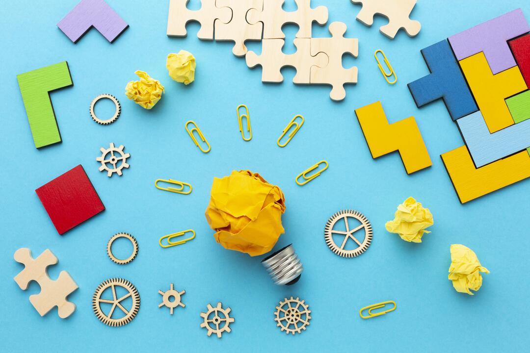 fotografía con una bombilla en el centro hecha de papel , piezas de rompecabezas, engranajes y fichas, como referencia a juagos de pensar.